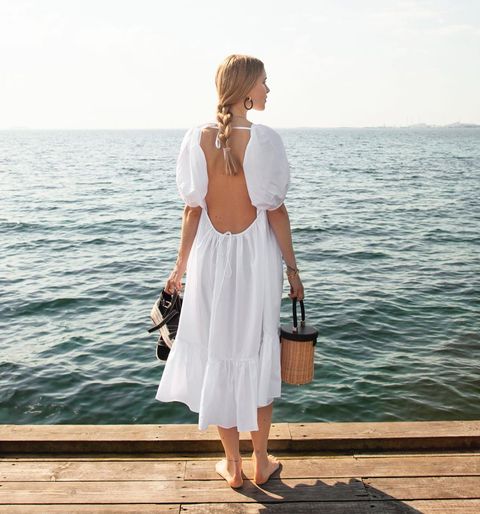 女孩身穿白色露背连衣裙，带着草编包和草编凉鞋站在海边