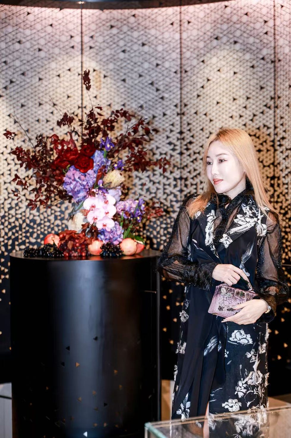 a person holding a stylish purse in a floral blouse next to a decorative display featuring flowers and fruits