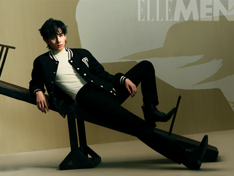male model posed on an inclined prop wearing a varsity jacket and black pants