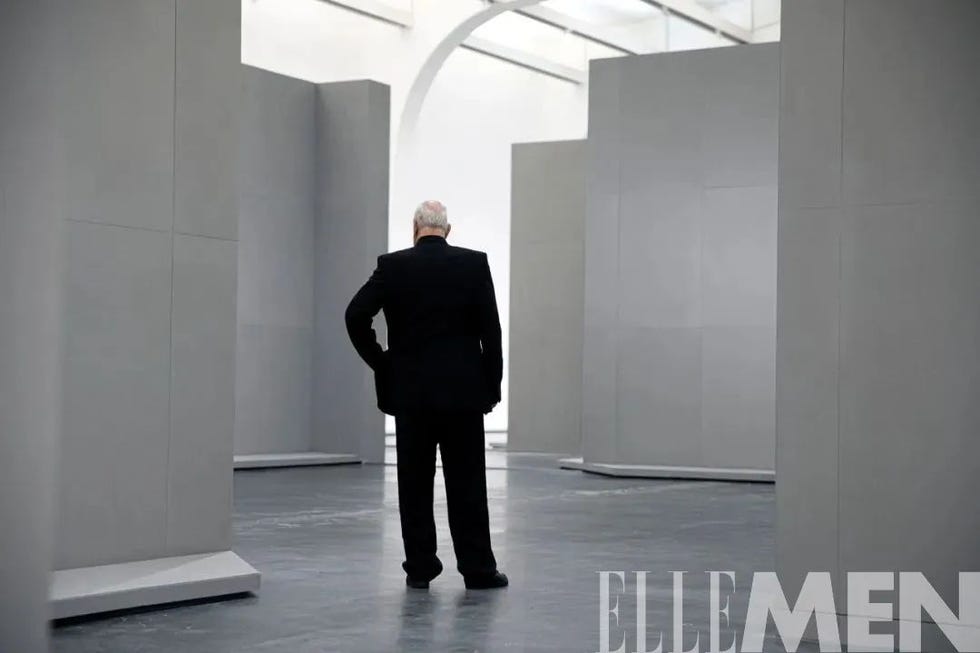 a man in a black suit stands in a gallery with tall gray partitions