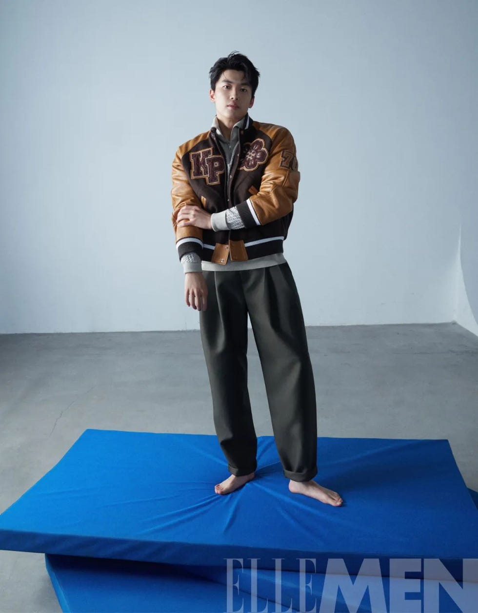 a model stands on blue mats wearing a brown varsitystyle jacket and gray trousers