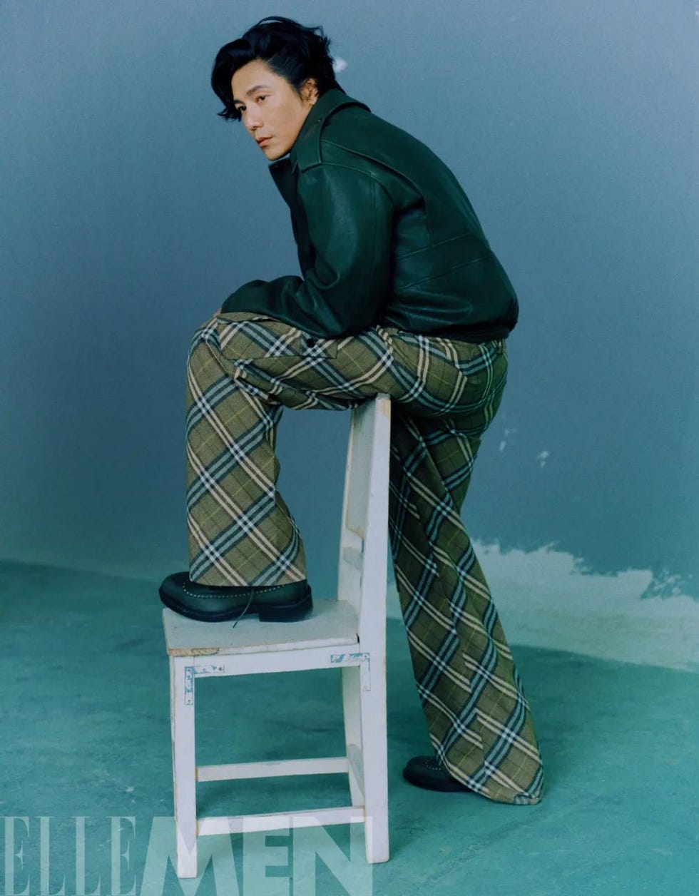 a person sits on a wooden chair wearing a leather jacket and plaid pants