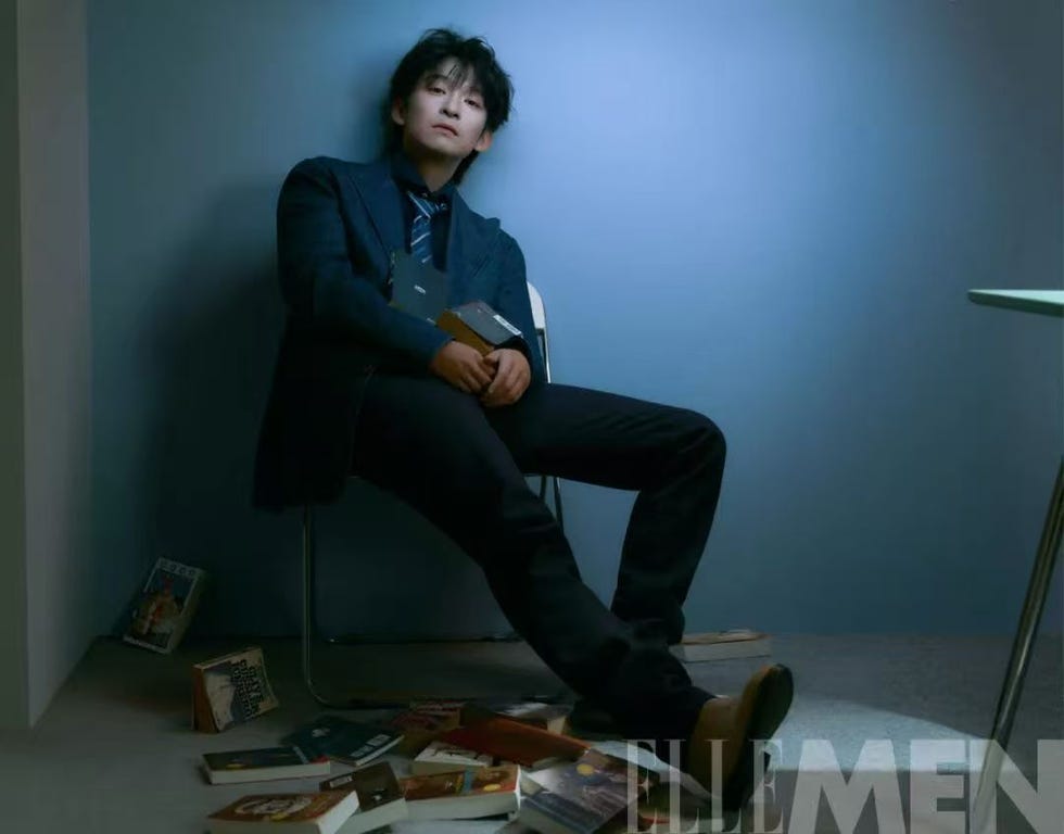 a seated person in a suit surrounded by scattered books in a minimalistic room
