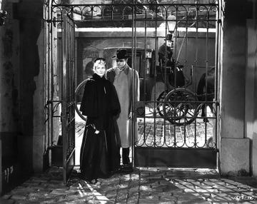 a man and a woman stand at a wrought iron gate with a horsedrawn carriage in the background