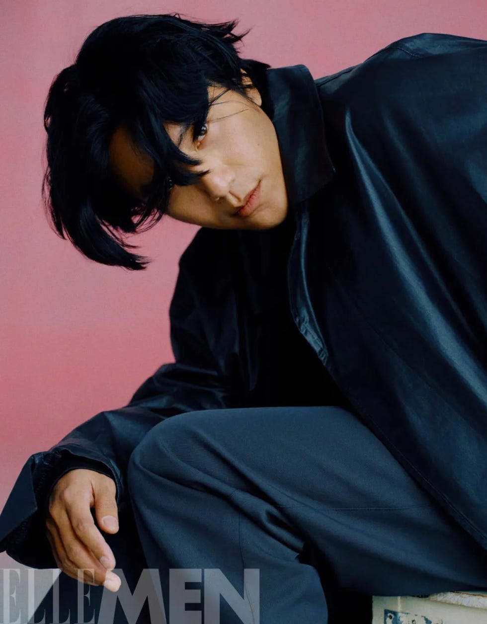 model seated in a dramatic pose against a pink background wearing a black oversized jacket and dark pants