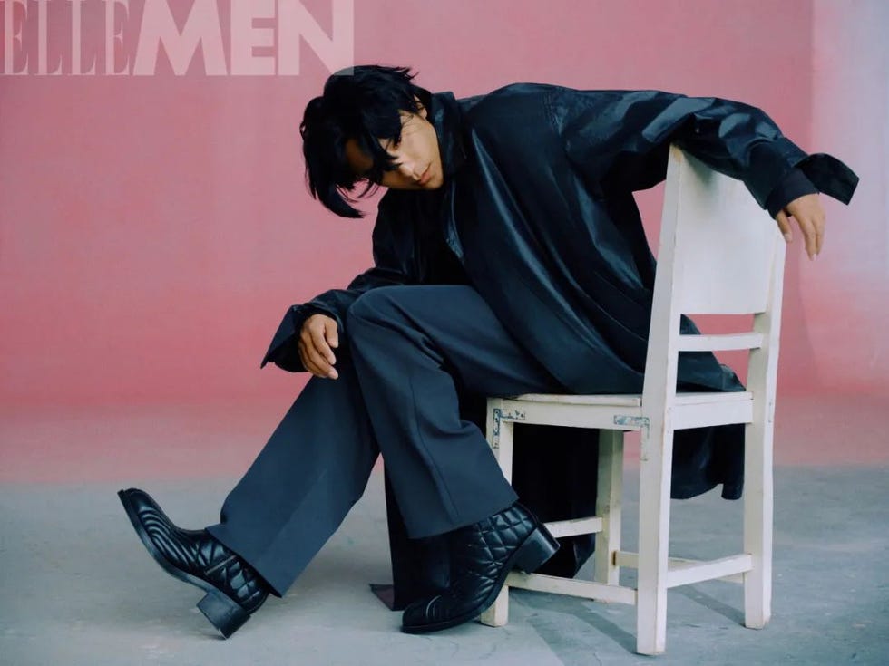 person in a black outfit seated on a white chair against a pink background