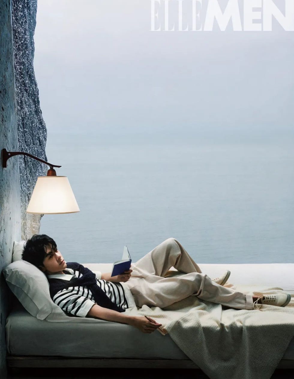 person relaxing on a bed with a book near a large window overlooking the sea
