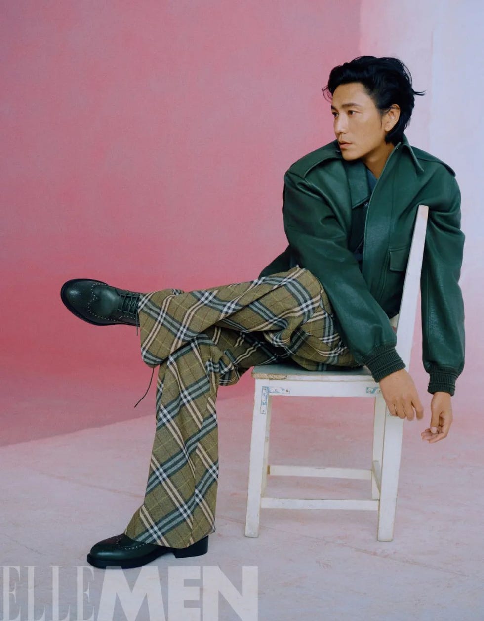 a seated model wearing a green leather jacket and plaid trousers against a pink background