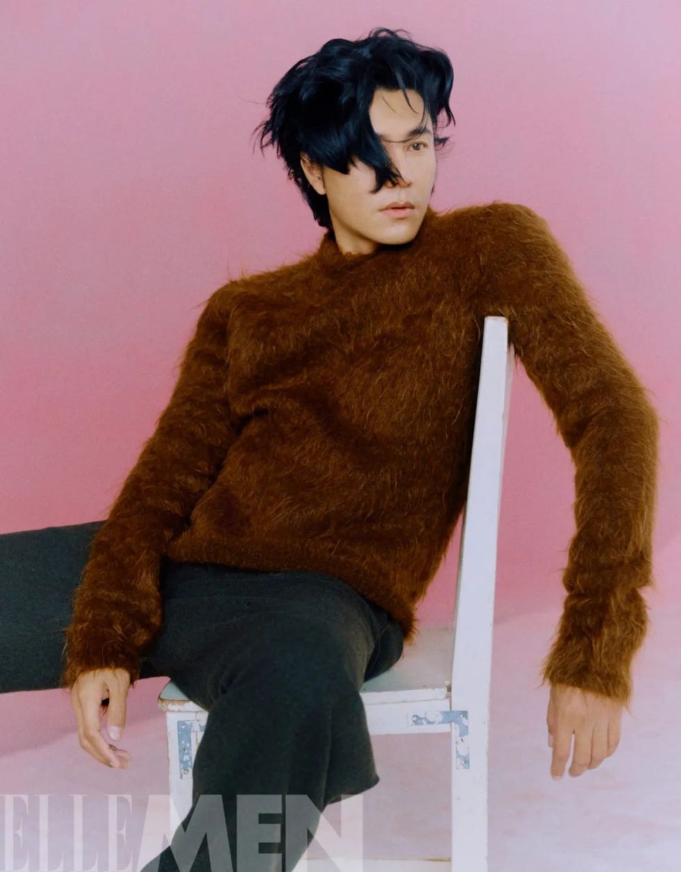 individual seated on a white chair against a pink background wearing a fuzzy brown sweater and dark pants