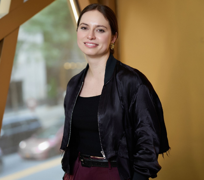person standing indoors wearing a bomber jacket and a black top with city scenery in the background