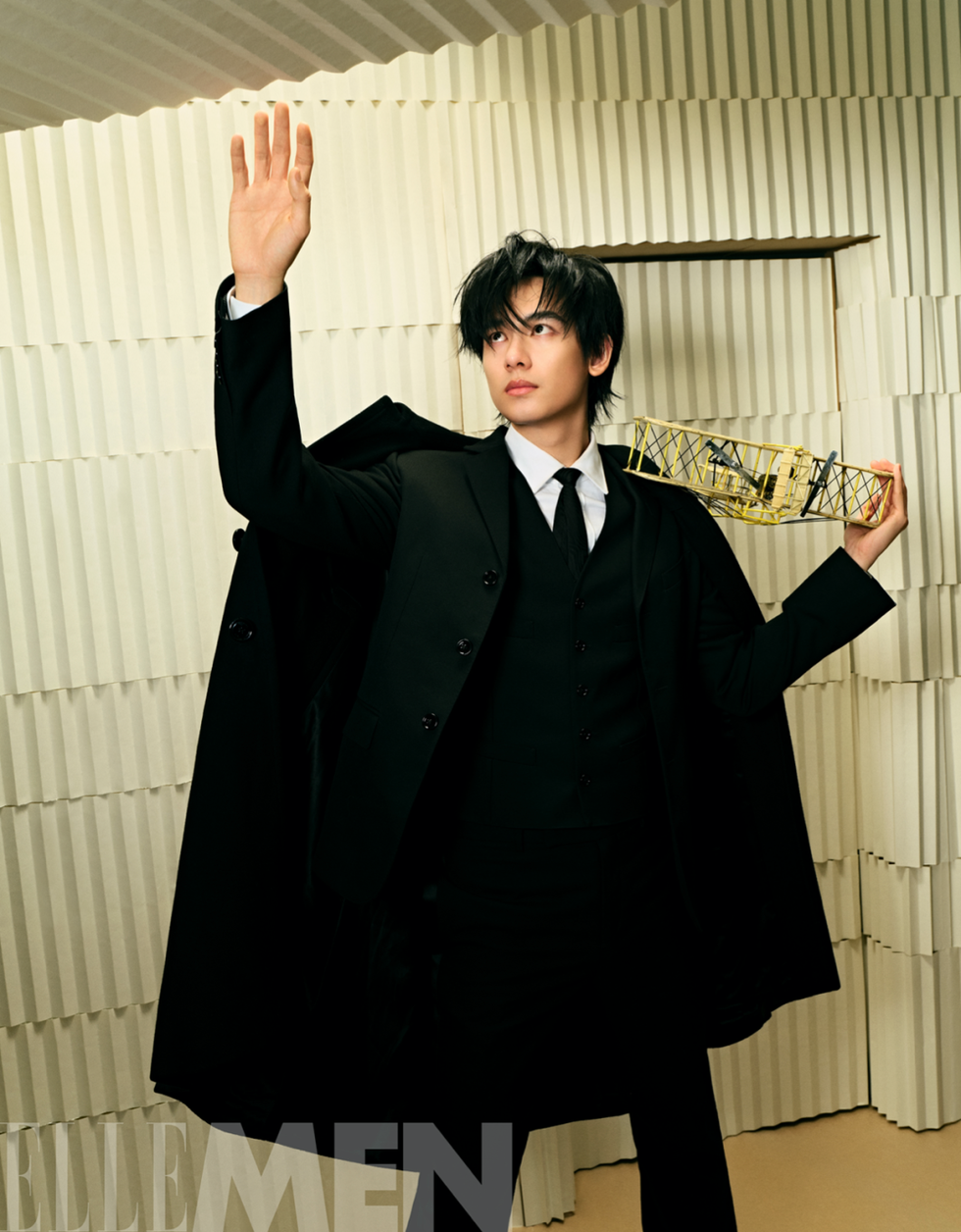 person in a formal black suit and long coat raising their hand and holding a yellow object