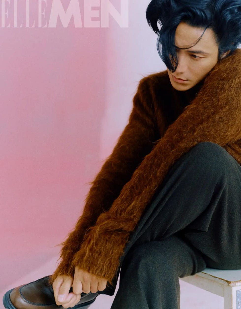 a seated figure with an obscured face wearing a fuzzy brown sweater and dark pants against a pink background