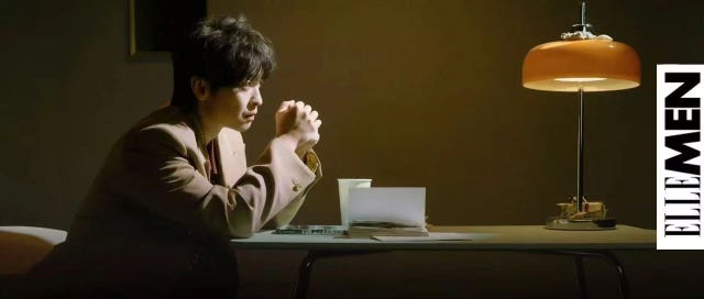man sitting at a table with folded hands beside a lamp and a stack of papers
