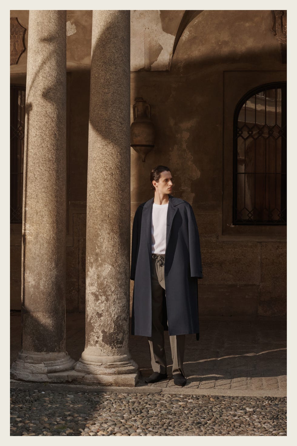 person in a long coat standing between two ancient columns in a rustic setting