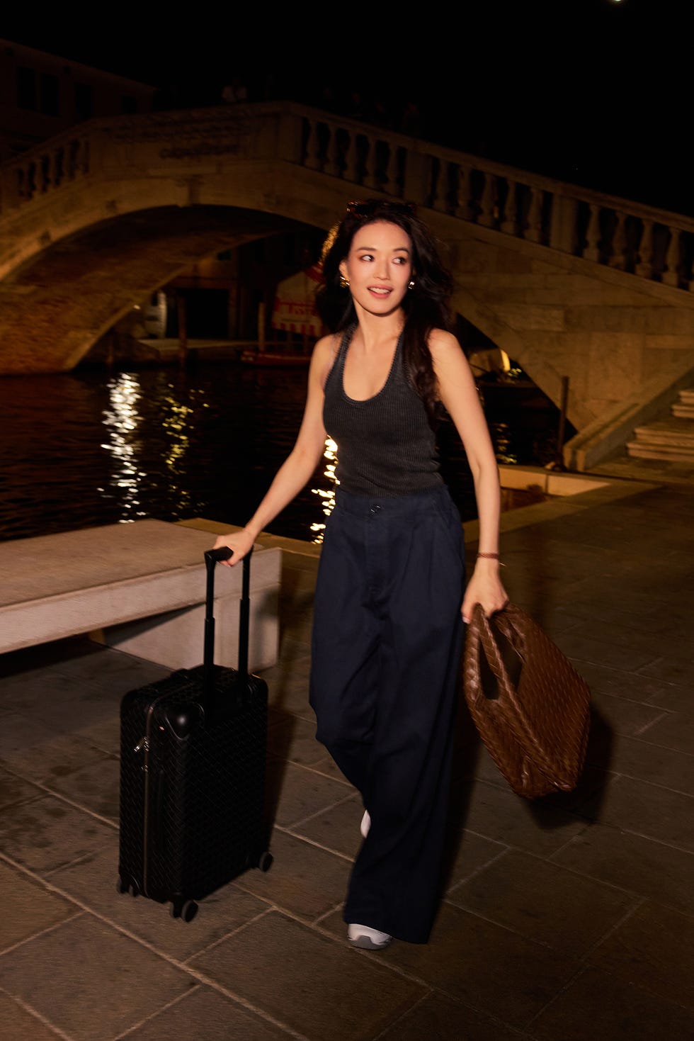 woman walking along a waterfront while carrying a suitcase and a bag