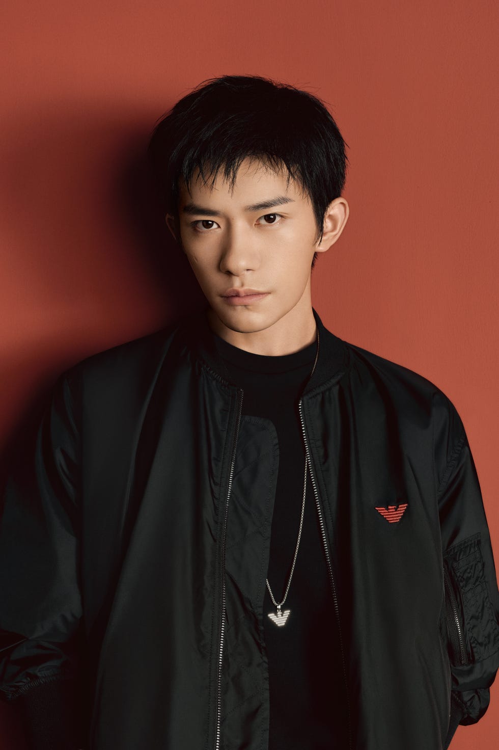 person in a black bomber jacket and necklace against a red background