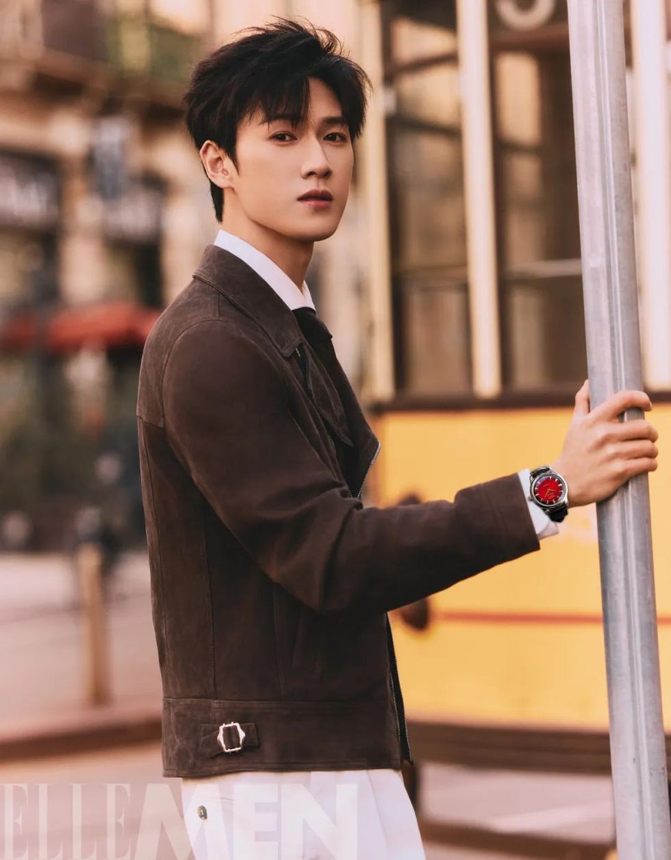 a man casually dressed in a brown suede jacket and white pants stands next to a pole