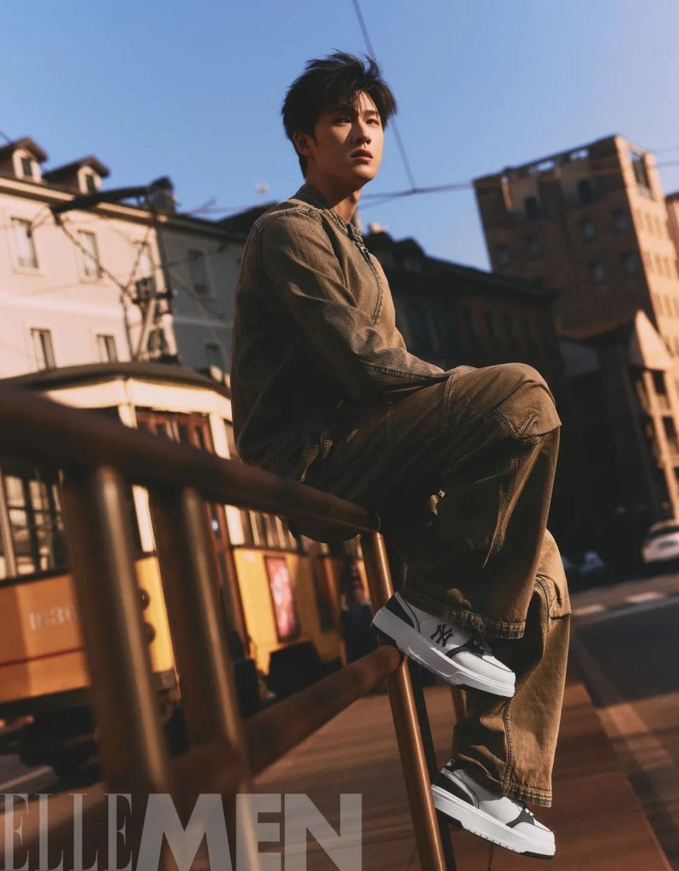 a person seated on a railing wearing stylish attire and sneakers in an urban setting during sunset