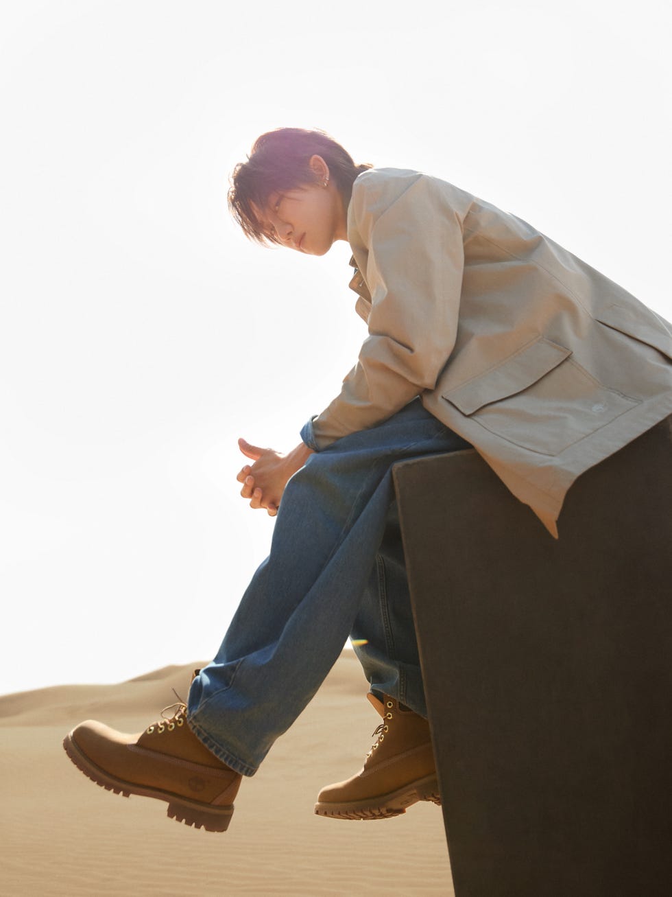 person wearing beige jacket and blue jeans sitting on a dark pedestal in a sandy environment