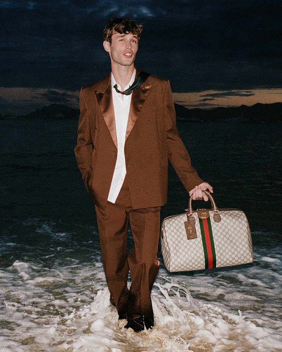 a person in formal attire standing in shallow water holding a designer bag
