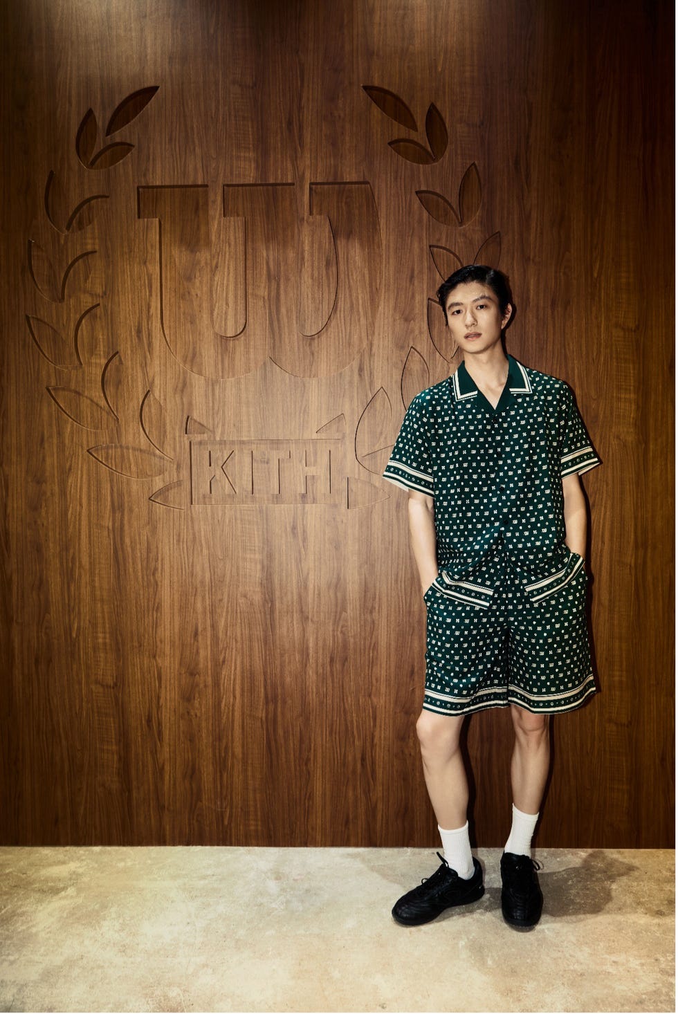 a person wearing a green patterned outfit stands against a wooden wall with a logo
