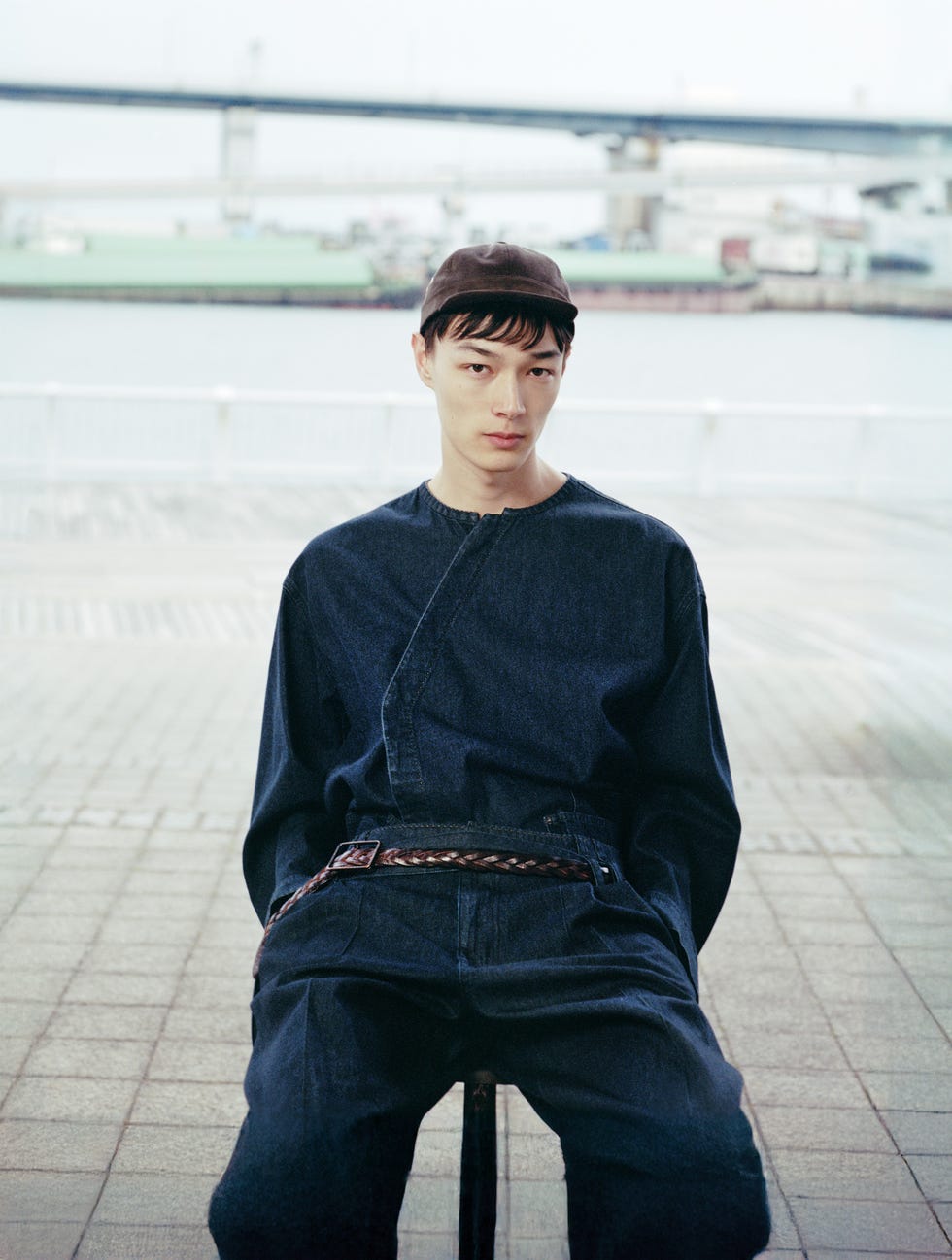 seated individual dressed in a dark denim outfit with a backdrop of a waterfront
